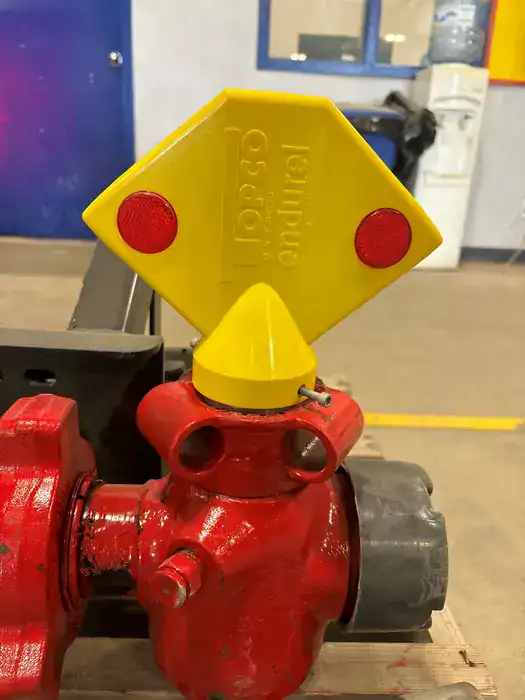 A close-up of a red industrial valve with a yellow diamond-shaped handle featuring two red circles and raised text. The background includes a blue door, window, and a water cooler in an indoor setting.