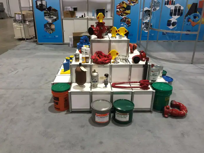 A display of industrial equipment and containers, including pipes, valves, paint cans, and protective gear, arranged on a stepped white platform at an exhibition booth with blue informational banners in the background.