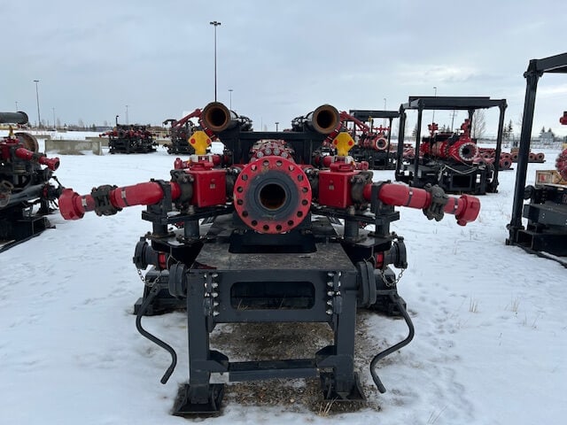 A large industrial valve assembly with red and black components sits on a metal frame outdoors in a snowy lot, surrounded by similar equipment under an overcast sky.