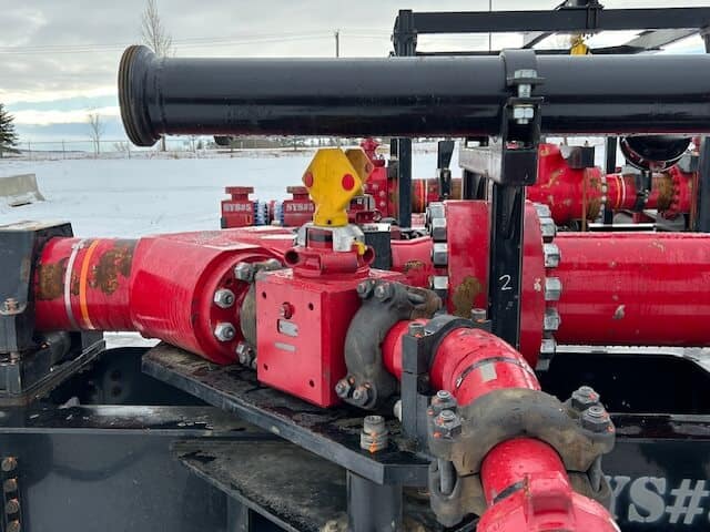 Close-up of red industrial pipes and valves connected together outdoors, with snow on the ground and a cloudy sky in the background. The equipment appears to be part of an oil or gas facility.