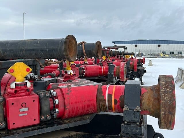 Rows of large red industrial pumps or pipes are lined up outdoors on a snow-covered surface, with a building in the background under a cloudy sky.