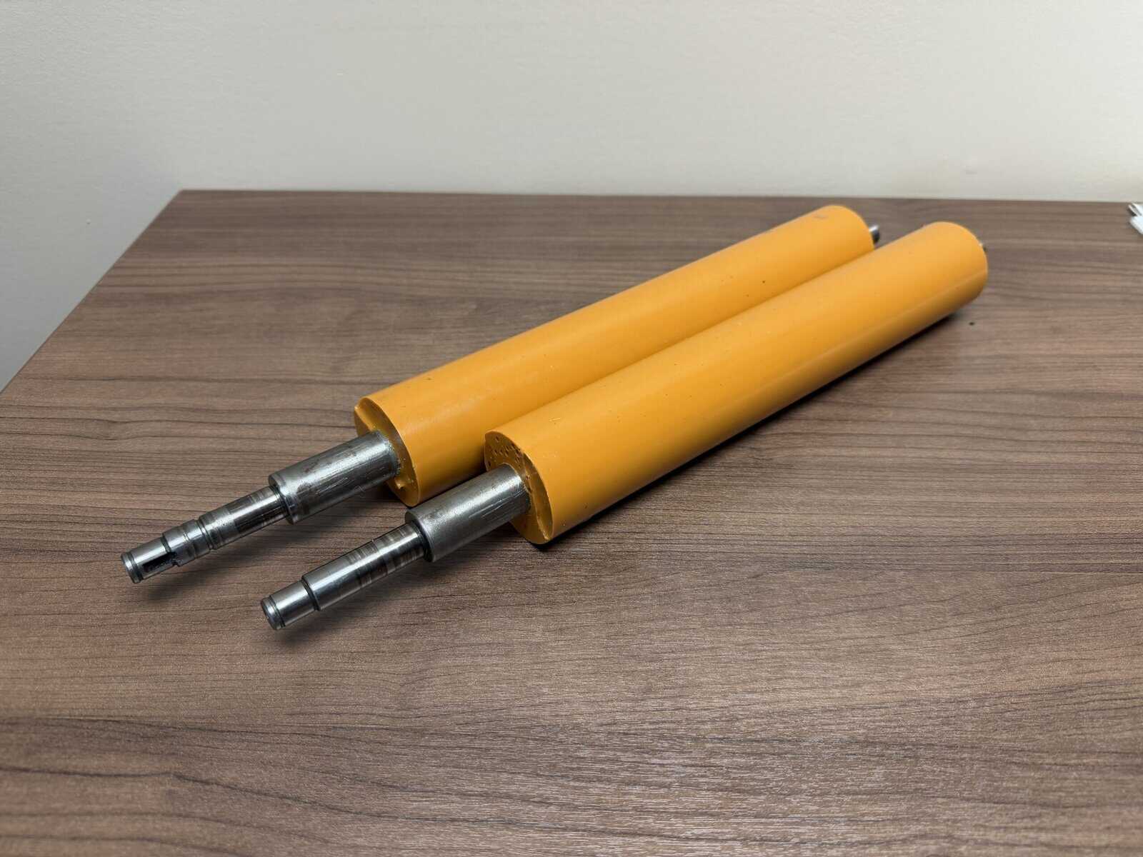 Two yellow cylindrical rollers with metal shafts on one end are placed side by side on a wooden surface.