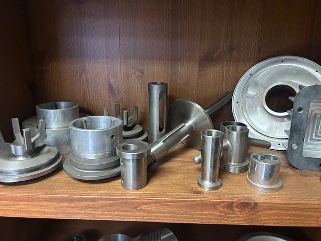 A collection of various machined metal parts, including cylindrical components and flanges, arranged on a wooden shelf against a wood-paneled background.