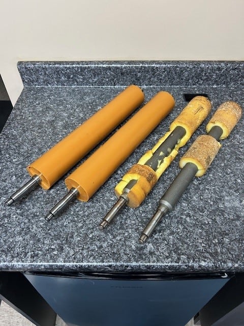 Four cylindrical rollers with metal shafts are placed on a speckled countertop; two rollers are intact with orange coverings, while the other two have torn and deteriorated yellow coverings, exposing the inner core.