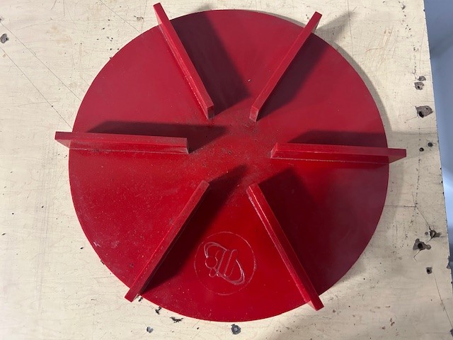 A round red metal object with six equally spaced upright fins on top, placed on a light-colored wooden surface. An engraved logo or initials are visible near the edge of the circle.